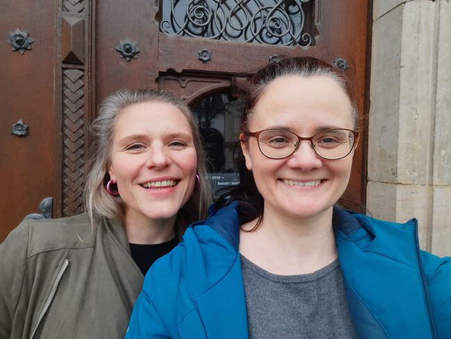 Levke und Toni (rechts) stehen lachend vor einer Holztür.